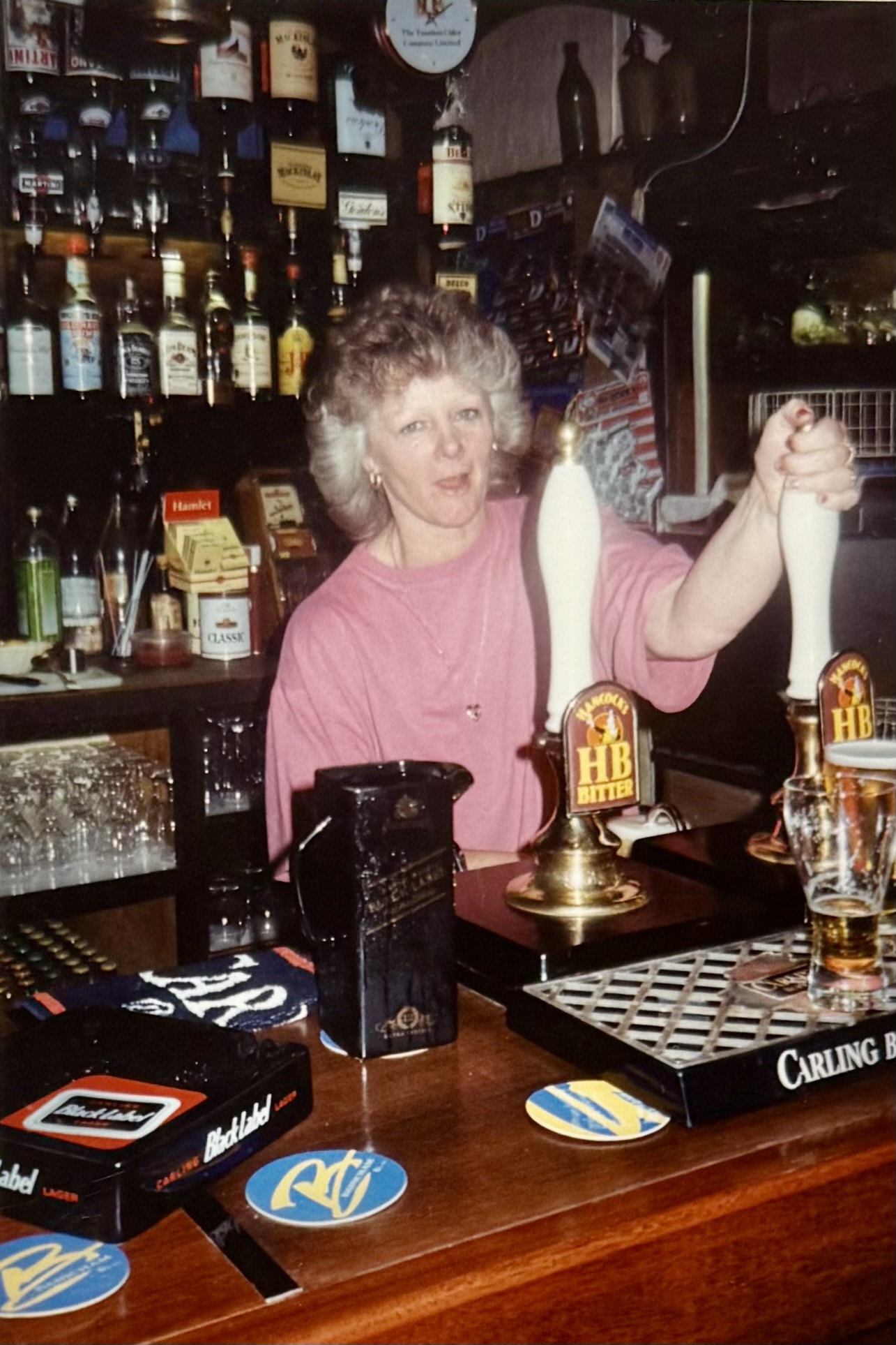 Anne, the barmaid at the time, standing behind the bar, pulling a pint of Hancock's Home Brew. You can see Hamlet cigars for sale behind her.