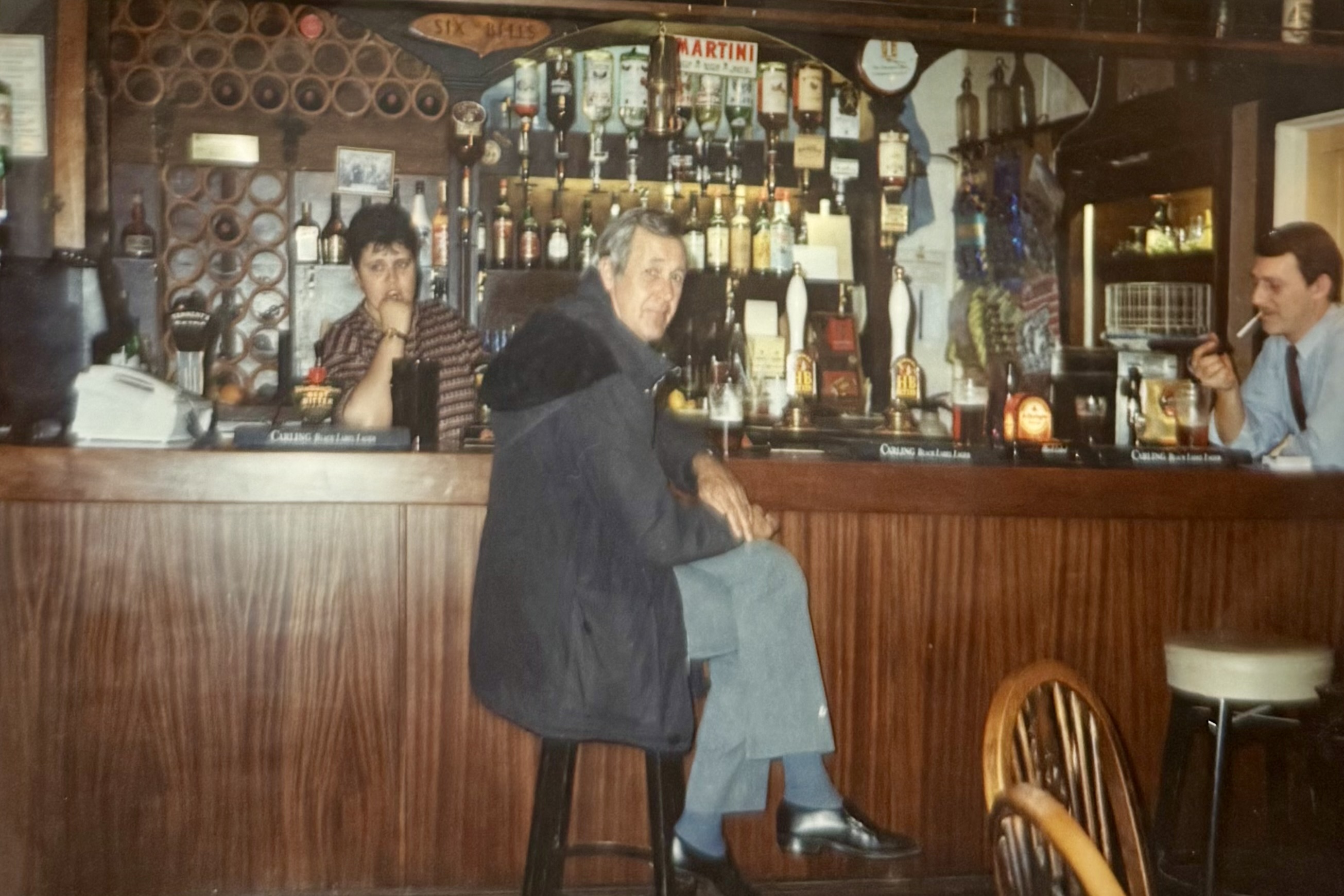 The bar, photo taken from what is now the Bar side. The photo shows a woman behind the bar, and two men at the bar, one of the smoking a cigarette.