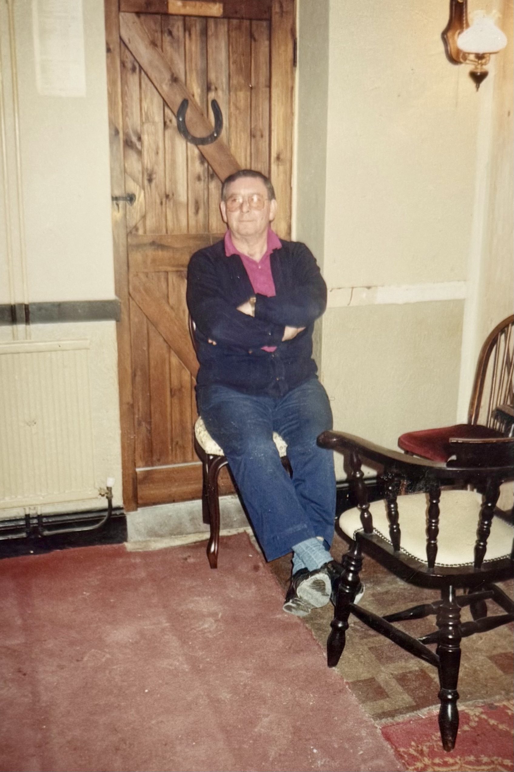 A man sat on a bar chair, in front of a wooden door with a horseshoe on it.
