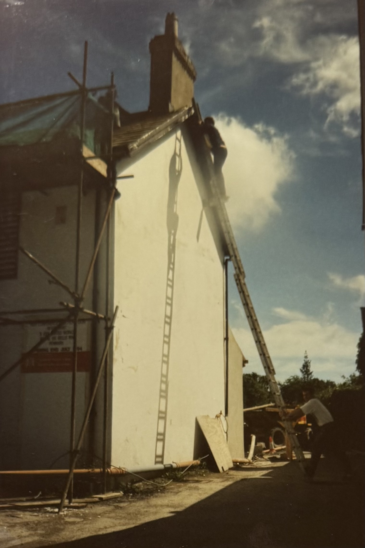 A man stood at the top of a very tall ladder, doing some work on the roof at the side of the pub. A man stands at the bottom holding the ladder.