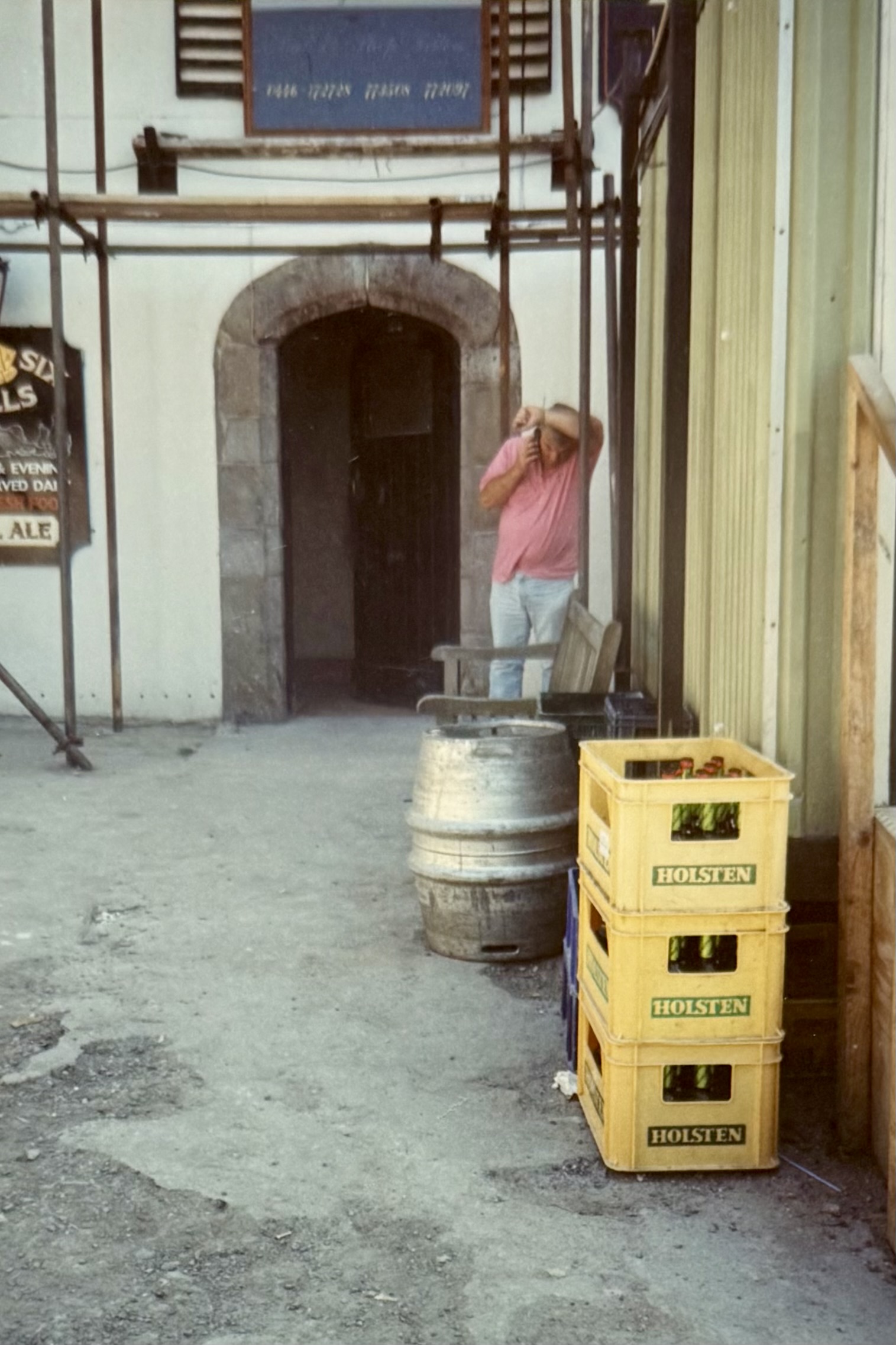 A man on the phone outside the front door of the pub. There's scaffolding behind him, a portakabin to the side, and some cases of beer and a barrel nearby.