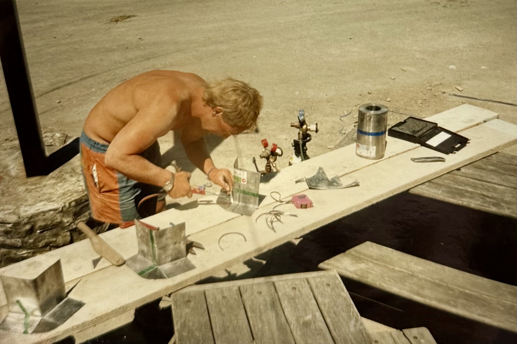 A man doing some work on a makeshift workbench, two planks of wood laying across some picnic benches.