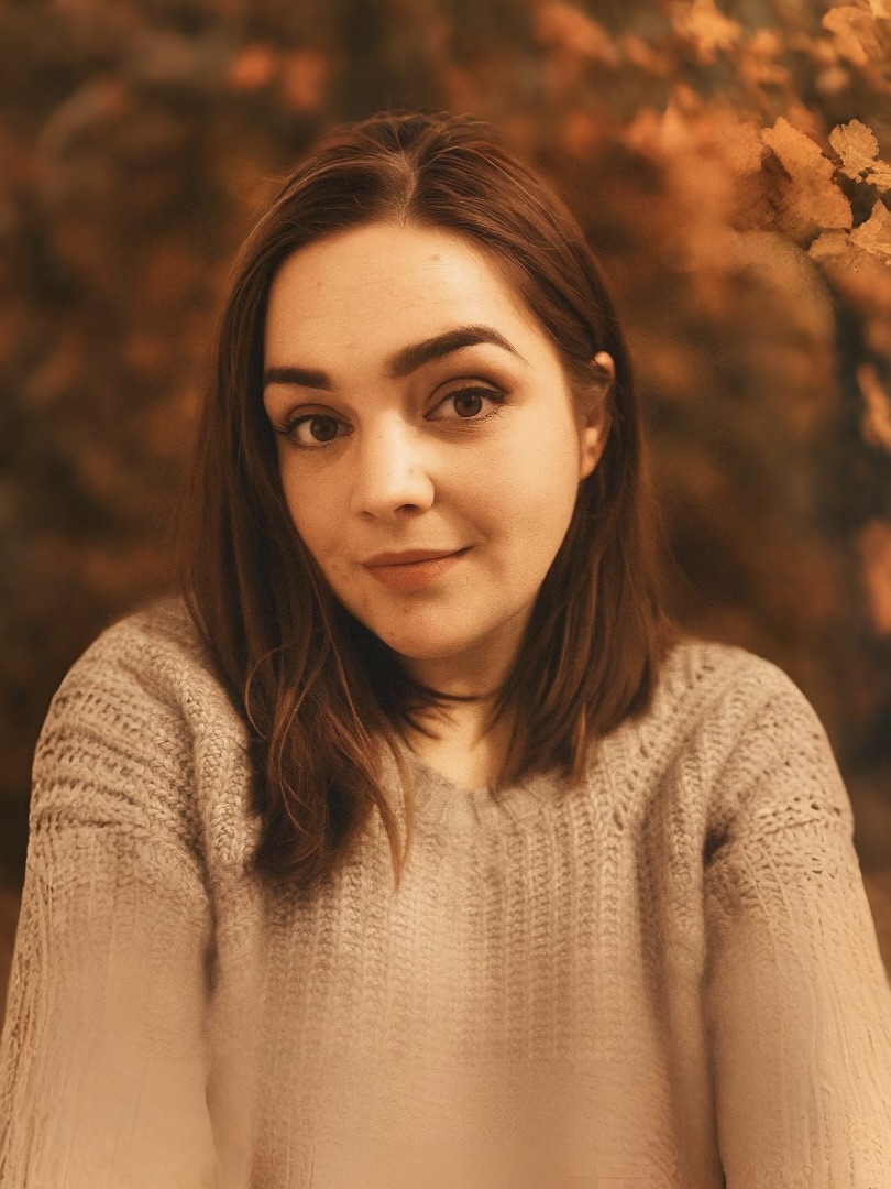 Becky wearing a grey jumper with an Autumnal background.